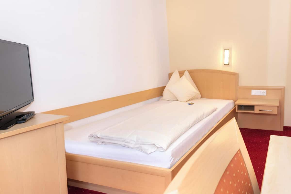 A hotel room with a single bed, a bedside table with a TV, a small bedside table with a lamp, and a red carpeted floor.