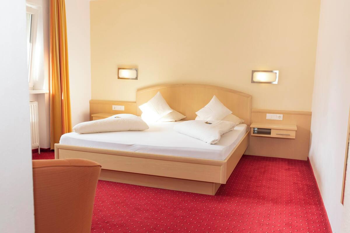 A hotel room with a bed, two bedside tables, a nightstand, and a red carpeted floor.