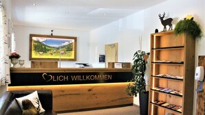 Ein Empfangsschalter mit Holzoptik und einem Willkommensschild, ein an der Wand befestigter Fernseher, ein schwarzes Ledersofa mit einem Zierkissen sowie ein Bücherregal mit Büchern und einer Pflanze.