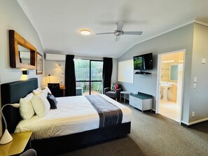 A modern hotel room with a large bed, a desk, a TV, and a balcony with a view of greenery.