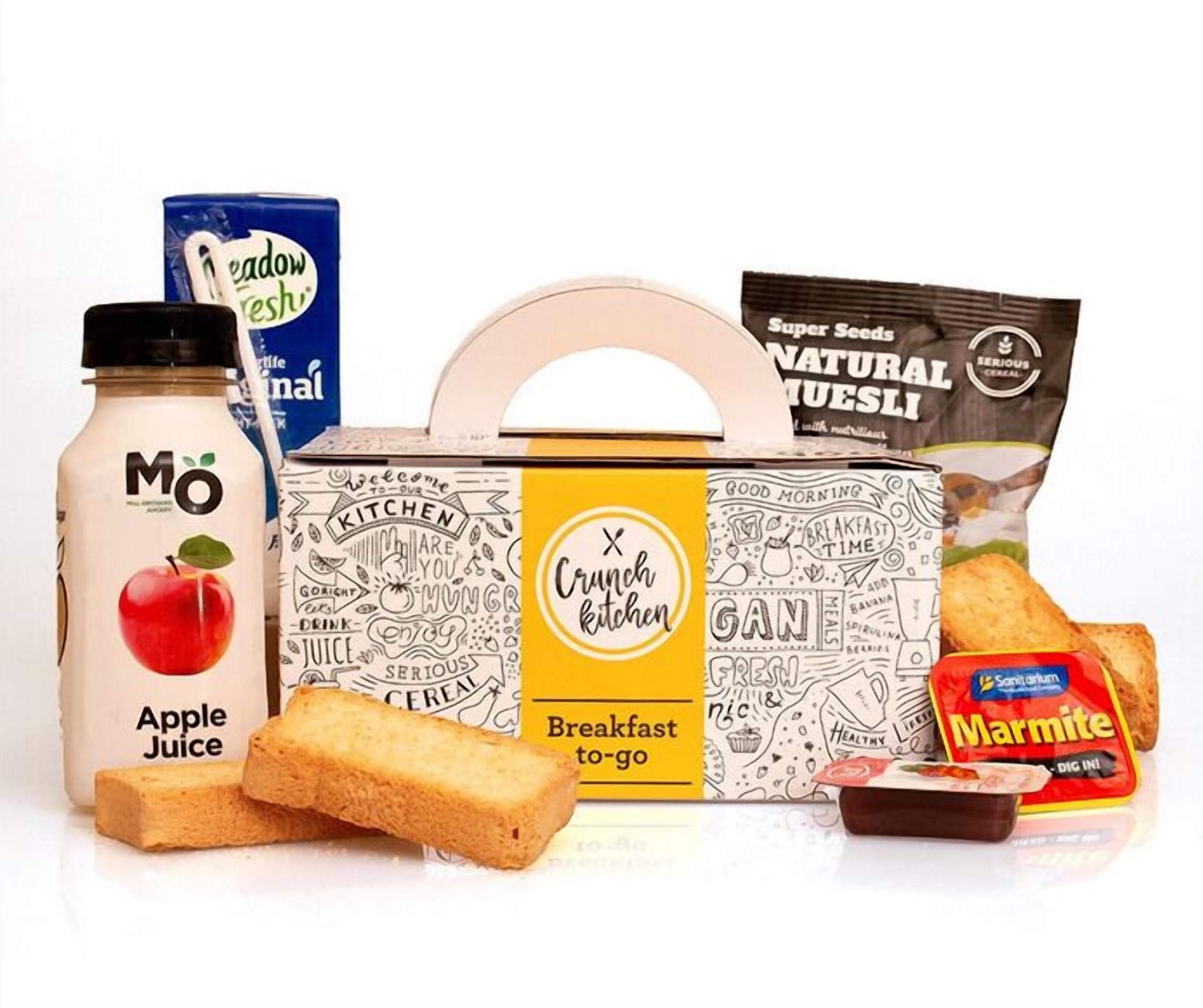A breakfast setup with a bottle of apple juice, a box of cereal, a bag of marmite, a bread roll, and a packet of super seeds.