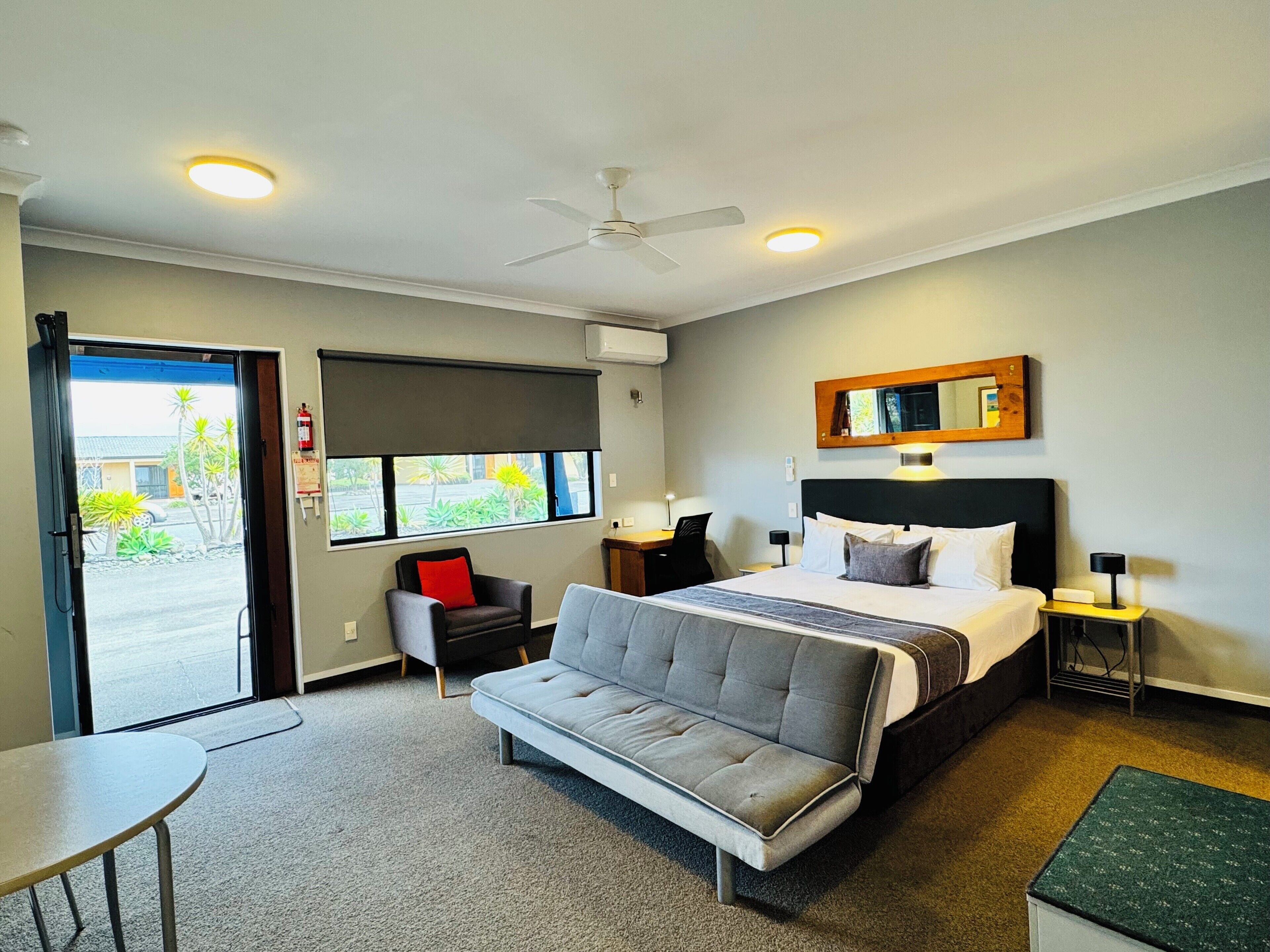 Hotelkamer met een bed, een slaapbank, een bureau, een stoel, een plafondventilator, een raam met uitzicht en een nachtkastje met een lamp.