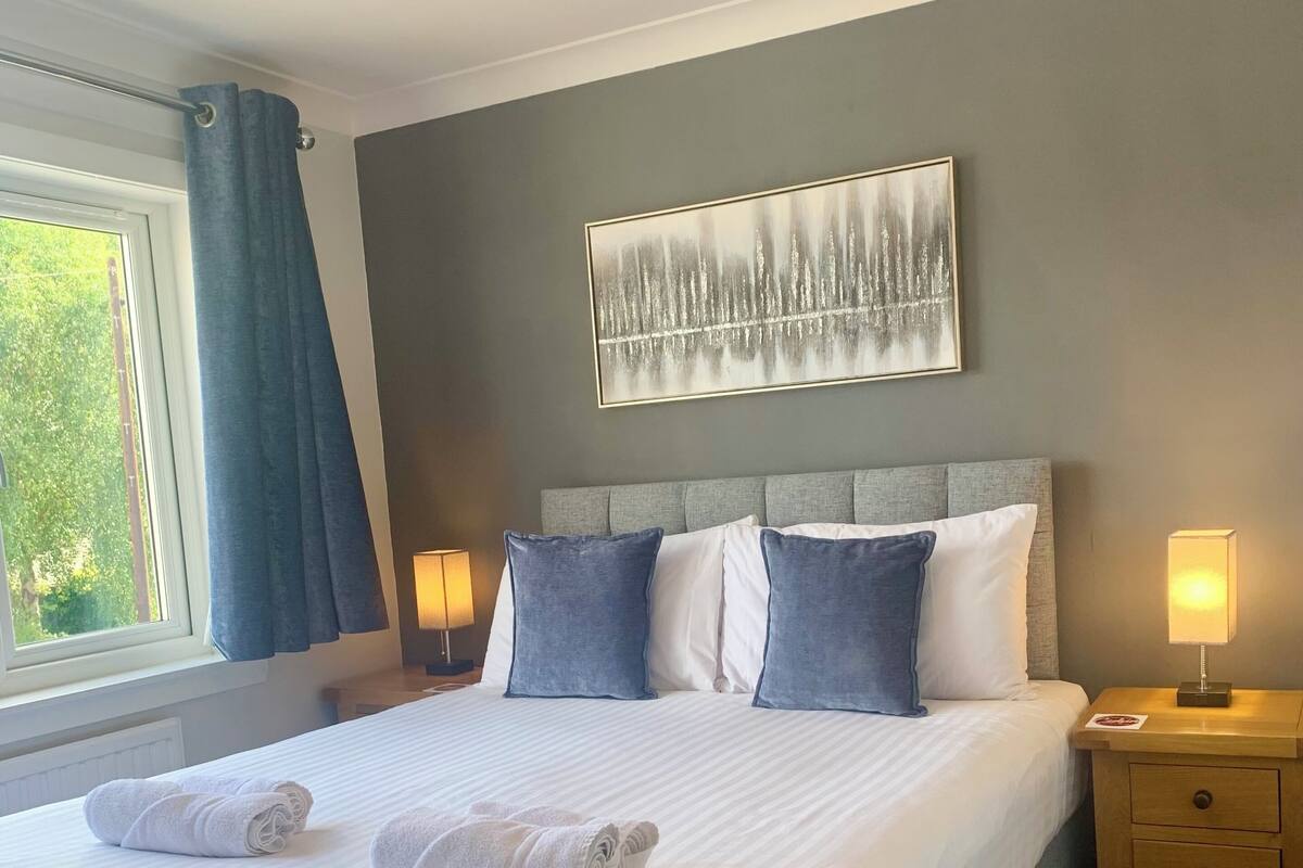 Uma cama bem arrumada com lençóis brancos e duas toalhas enroladas, uma cabeceira cinza, um quadro abstrato na parede, abajures e uma janela com cortinas.