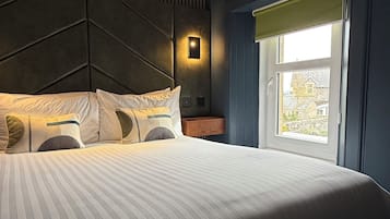 Una habitación de hotel moderna con una cama grande, cabecera con líneas verticales, mesita de noche y ventana con vista a edificios.