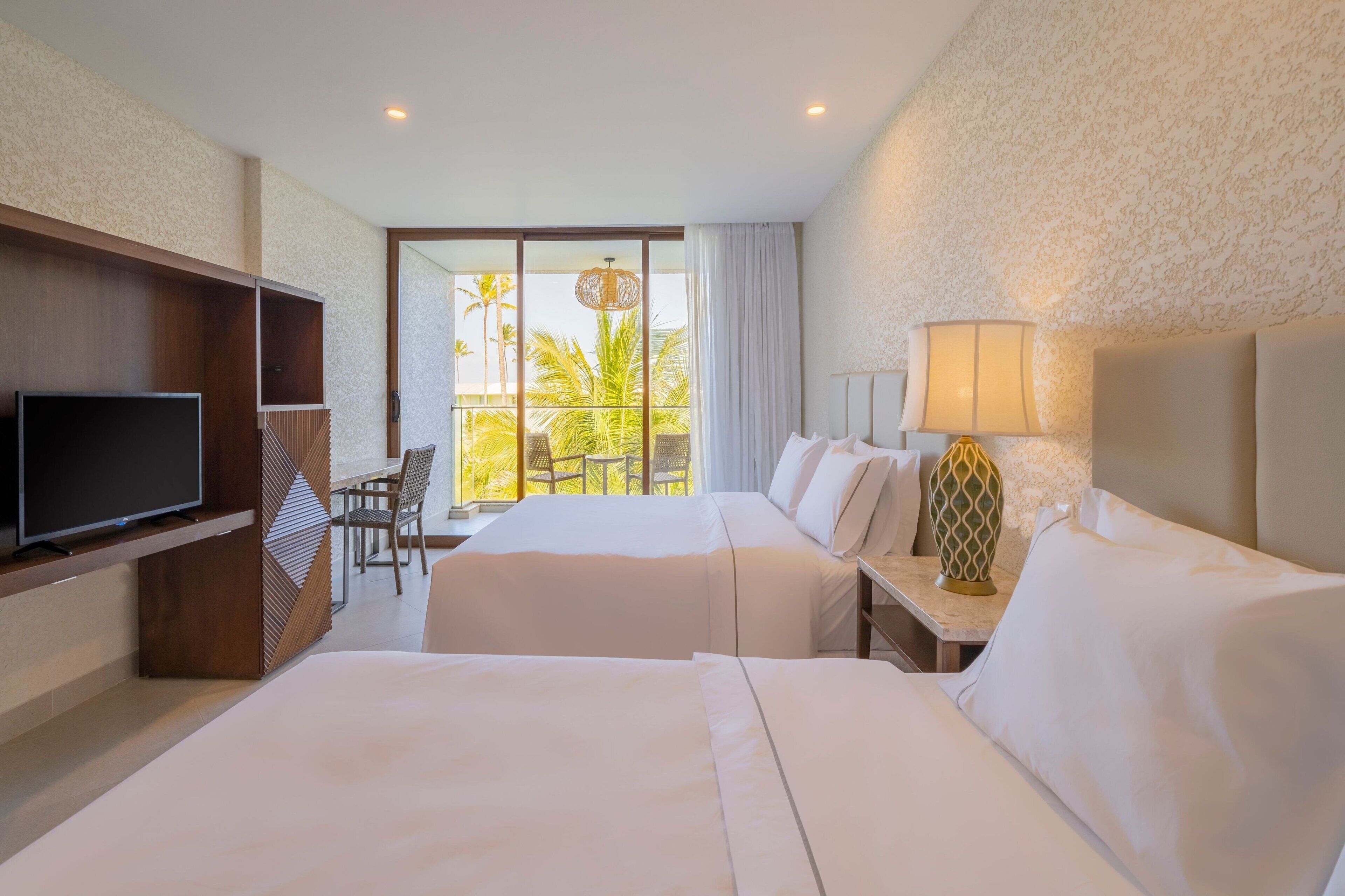A hotel room with two beds, a TV, a dining area with a table and chairs, and a balcony with a view of palm trees.