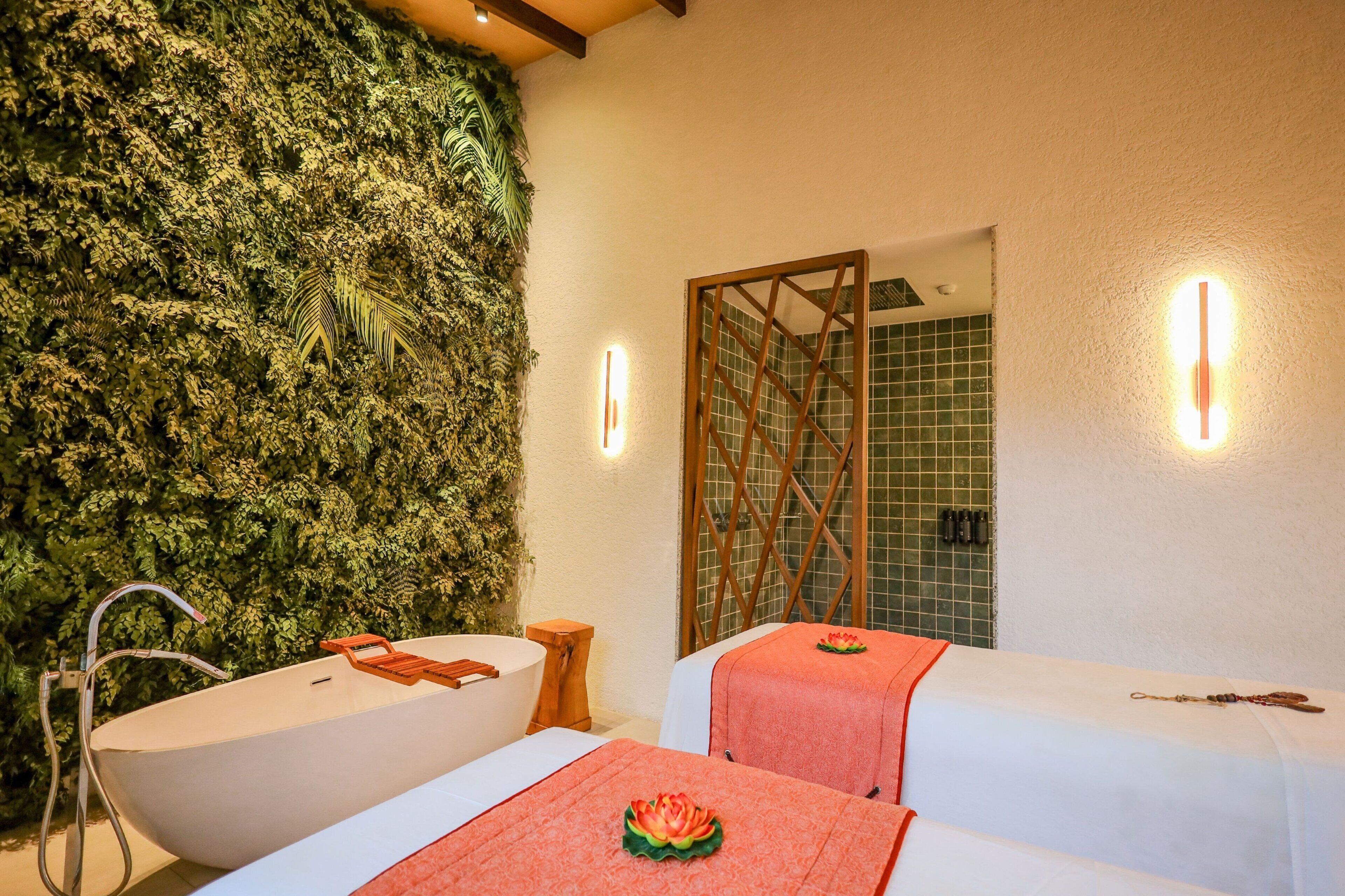 Un cuarto de spa con una bañera grande, dos camillas de masaje, un taburete de madera y una pared verde con follaje.