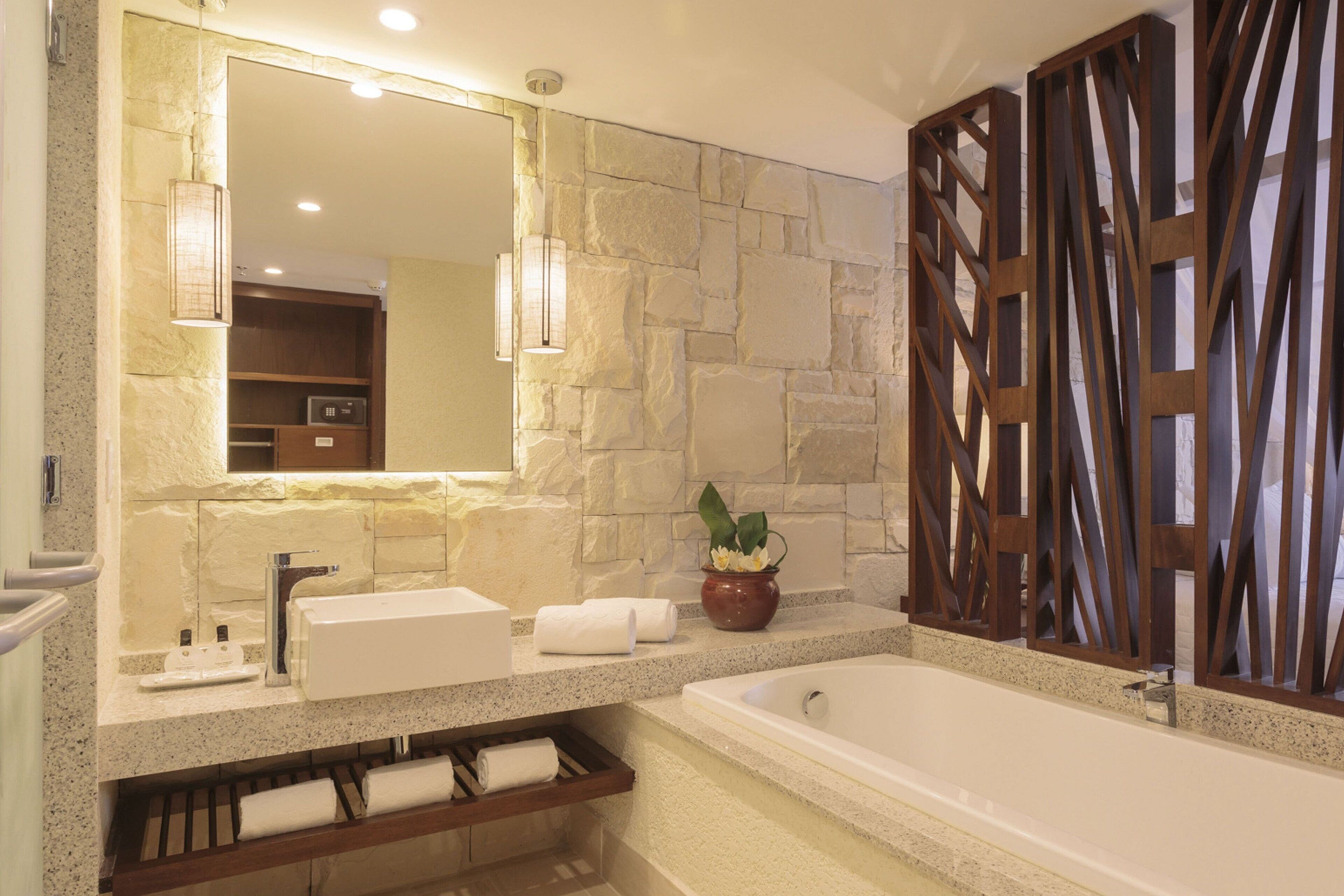 A bathroom with a stone wall, a large mirror, a sink, a bathtub, and a wooden partition.