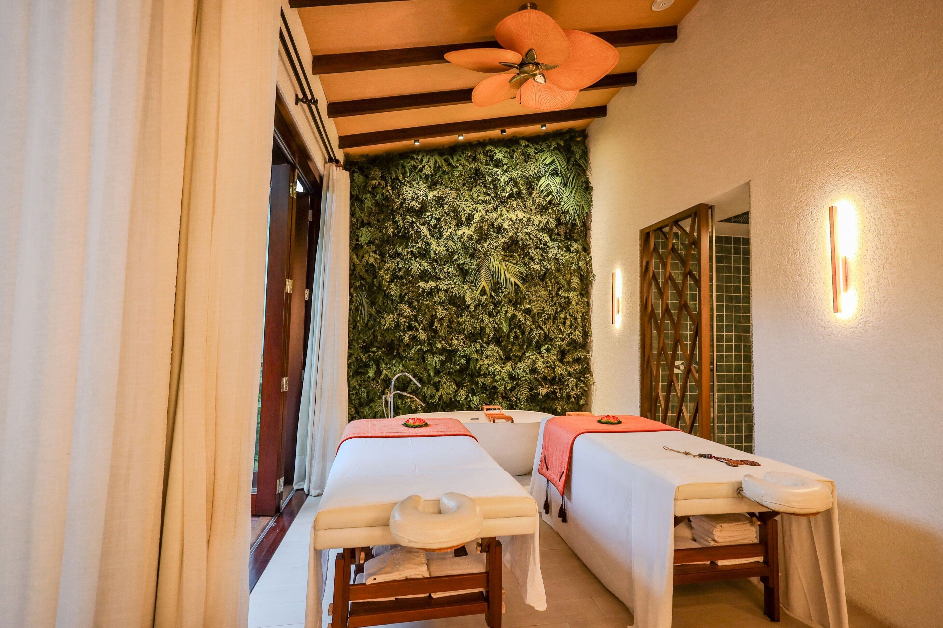 Sala de spa com duas mesas de tratamento, um ventilador de teto, uma parede com painel de folhagem e piso de azulejos.