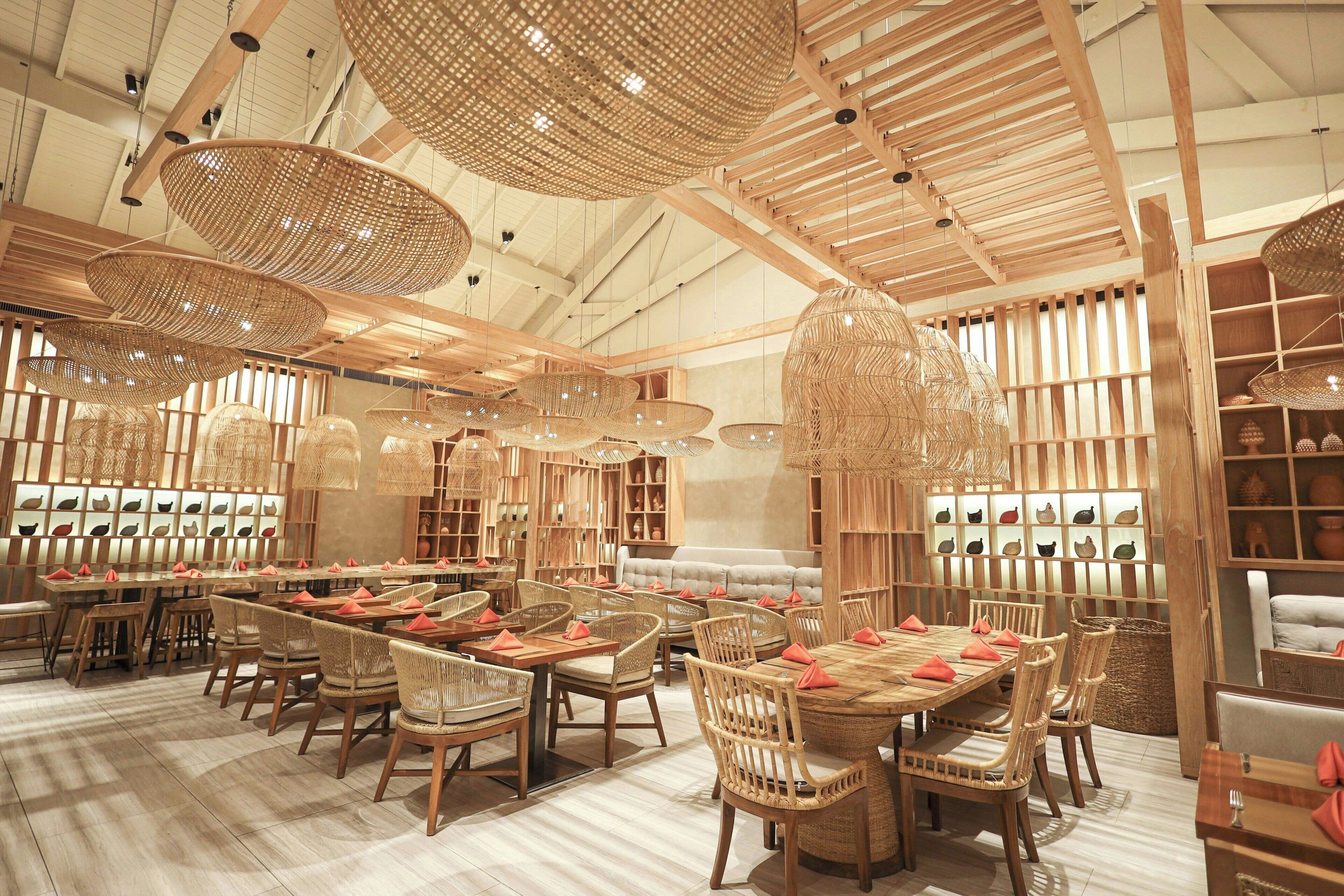 Ein geräumiges Restaurant mit hohen Decken, Holzbalken und gewobenen Lampen. Die Tische sind mit Korbstühlen und roten Servietten gedeckt.