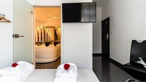 A hotel room with a bed, a TV mounted on the wall, a bathroom with a bathtub, and a seating area with a chair.