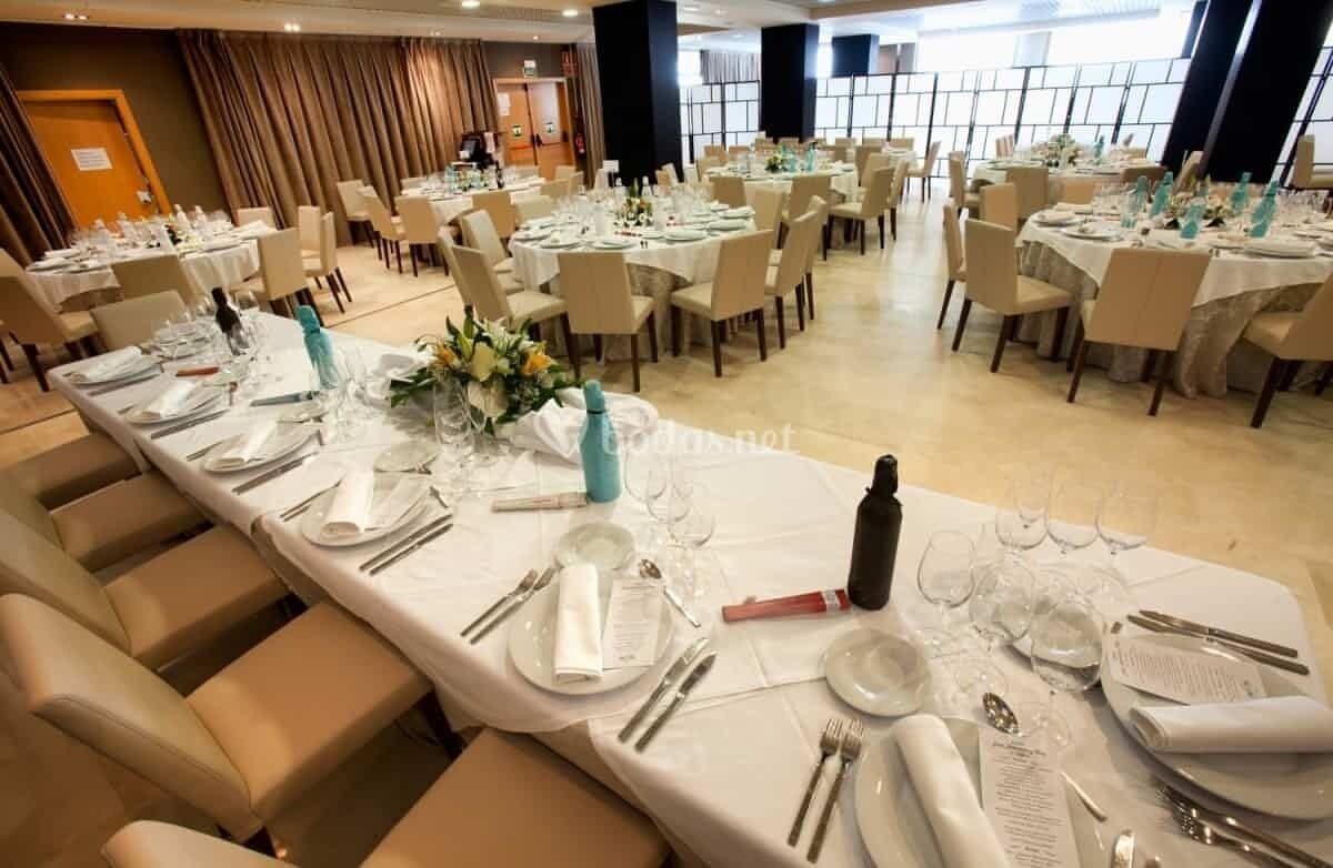 Une salle de banquet avec de nombreuses tables rondes dressées pour le repas, chacune étant recouverte d’une nappe blanche et agrémentée d’assiettes, de couverts et de verres.