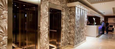 A modern hotel lobby with two elevators, a reception desk, and a decorative wall with a leaf pattern.