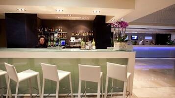 Una zona de bar con una barra larga, varios taburetes blancos y estantes llenos de botellas.