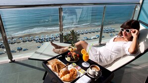 Iemand ontspant op een ligbed met een dienblad met ontbijtartikelen en een drankje, met uitzicht op een strand met parasols en mensen.