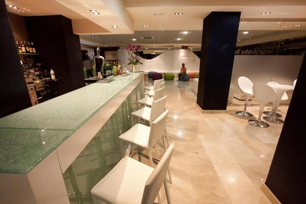 Zona de bar moderna con mostrador de vidrio, sillas blancas y un área de descanso con cojines de colores.