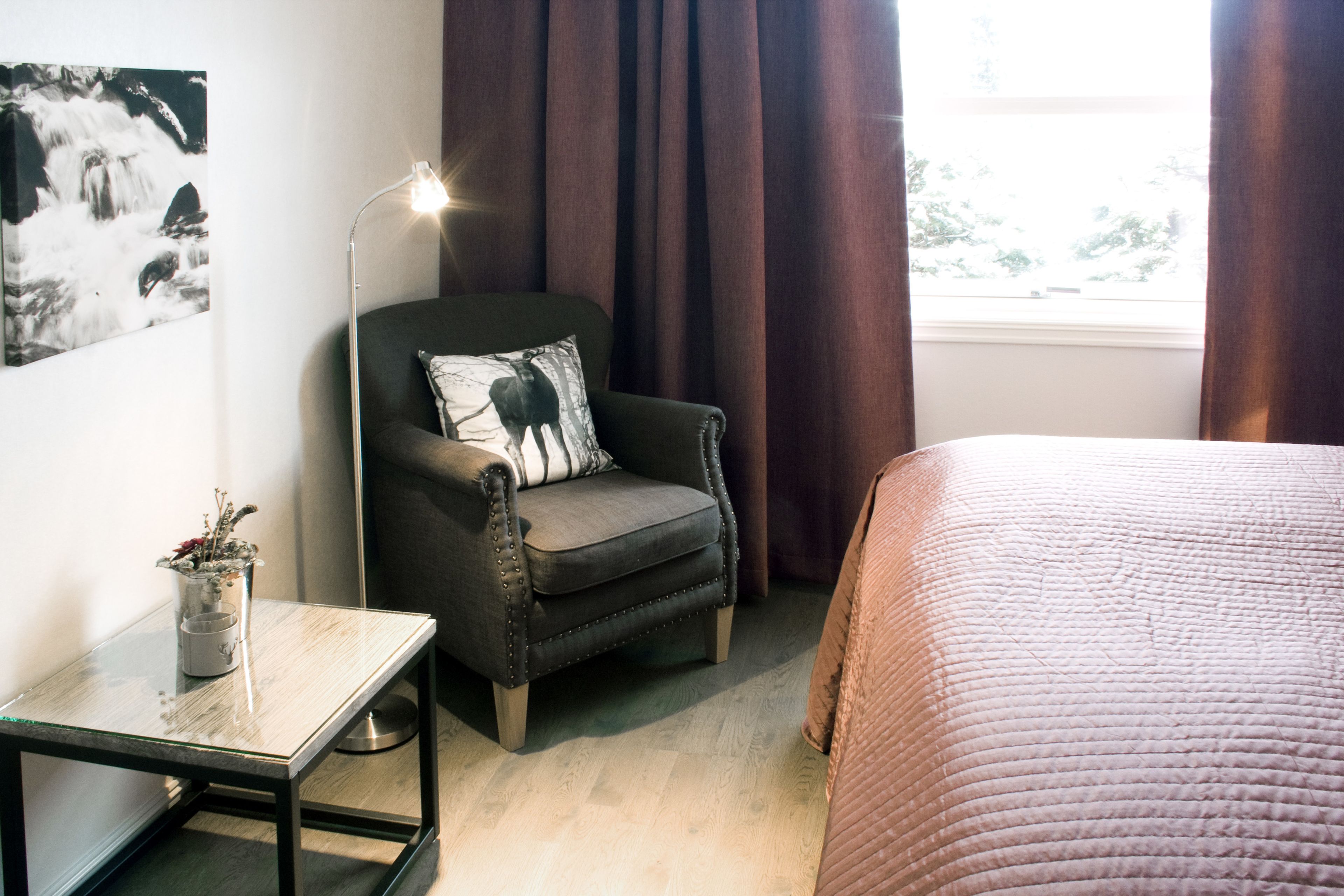 Una habitación con una cama, una silla con un cojín, una mesa con un florero y una lámpara de pie.