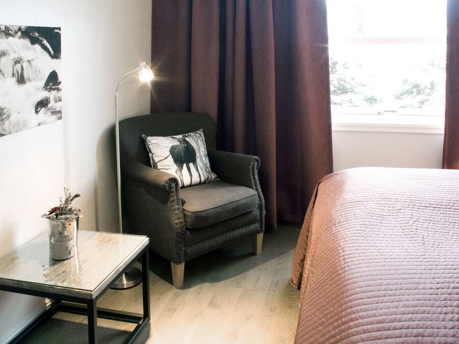 A room with a bed, a chair with a pillow, a table with a vase, and a floor lamp.