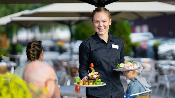 En tjener serverer mad i et udendørs spiseområde med borde og parasoller.