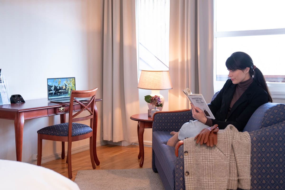 A hotel room with a wooden desk, a laptop, a chair, a sofa, a lamp, and a window with curtains.
