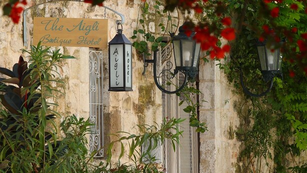 Un bâtiment ancien, sur lequel on peut lire l’inscription « The Aigli Boutique Hotel », agrémenté de plantes en pot et de lampadaires décoratifs.