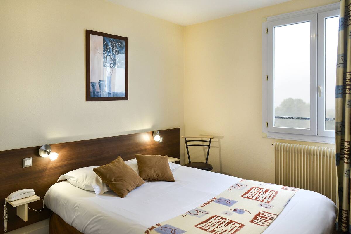 Habitación de hotel con cama, mesita de noche con teléfono, luz de pared, ventana con cortinas y radiador.