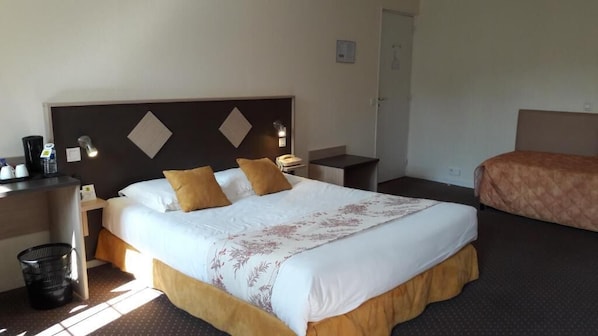 A hotel room with a large bed, a bedside table with a lamp, a desk with a chair, and a trash bin.