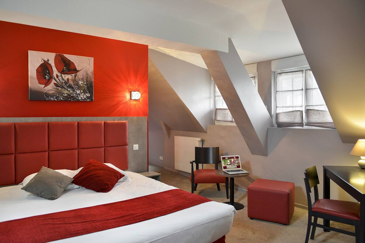 Una habitación de hotel moderna con una decoración en tonos rojo y blanco, una cama grande, un escritorio con una computadora portátil y vistas del cielo a través de un tragaluz.
