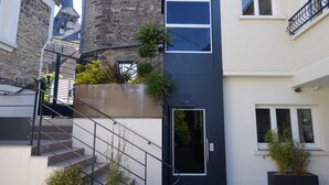 A modern building with a staircase leading to an entrance, a stone tower in the background, and potted plants near the entrance. - Au Site Normand (Clecy)