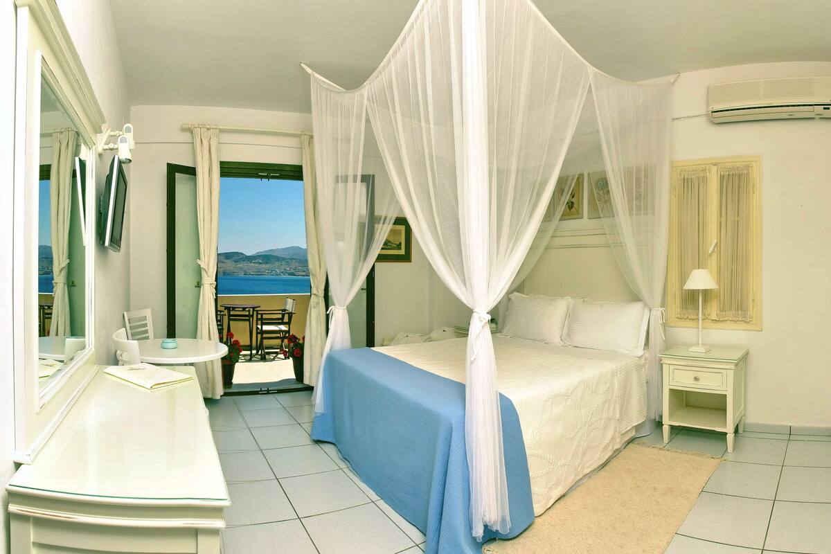 A bedroom with a bed, a bedside table, a nightstand, a mirror, a window with curtains, and a view of the sea.