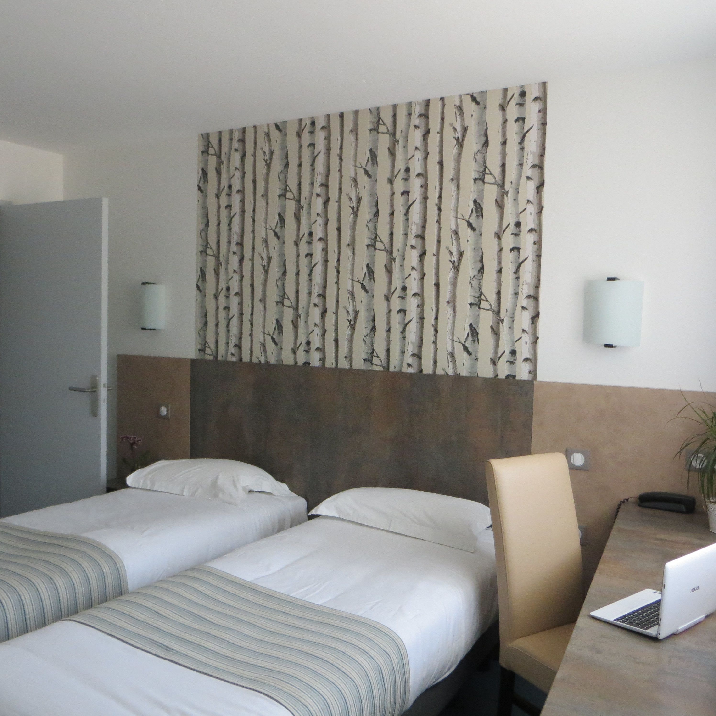A hotel room with two beds, a desk with a laptop, and a wall with a tree pattern.