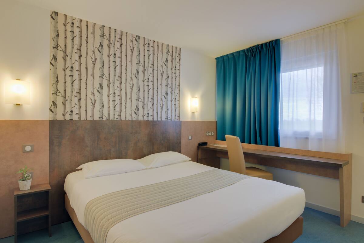 Une chambre d’hôtel comprenant un lit, un bureau, une chaise, un téléphone fixé au mur, une lampe fixée au mur et une fenêtre avec des rideaux.