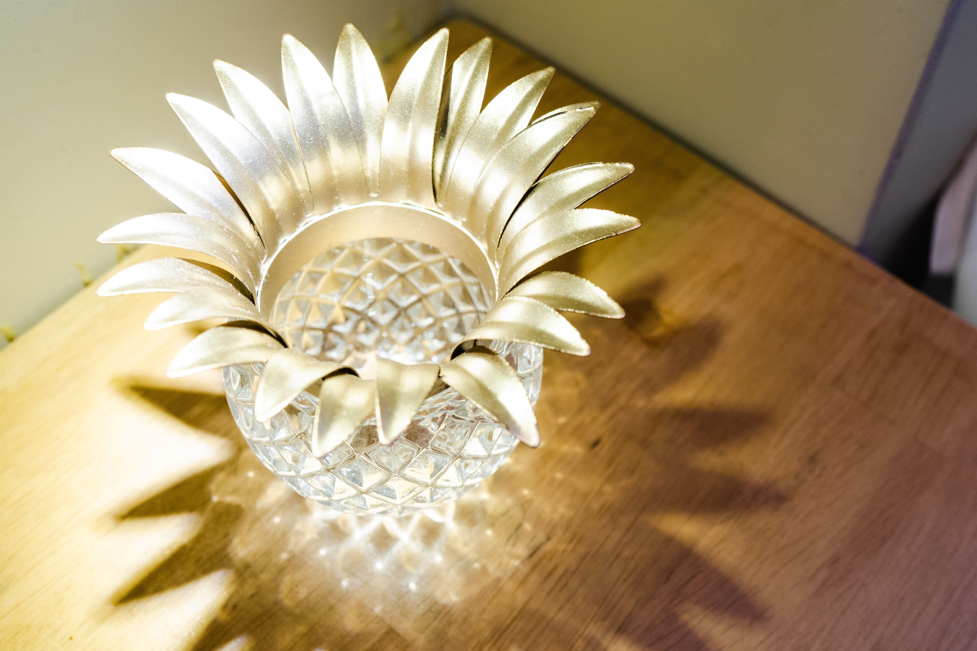 Een transparant glazen object met een geometrisch patroon en een metalen, bloembladachtige structuur bovenop, geplaatst op een houten ondergrond.