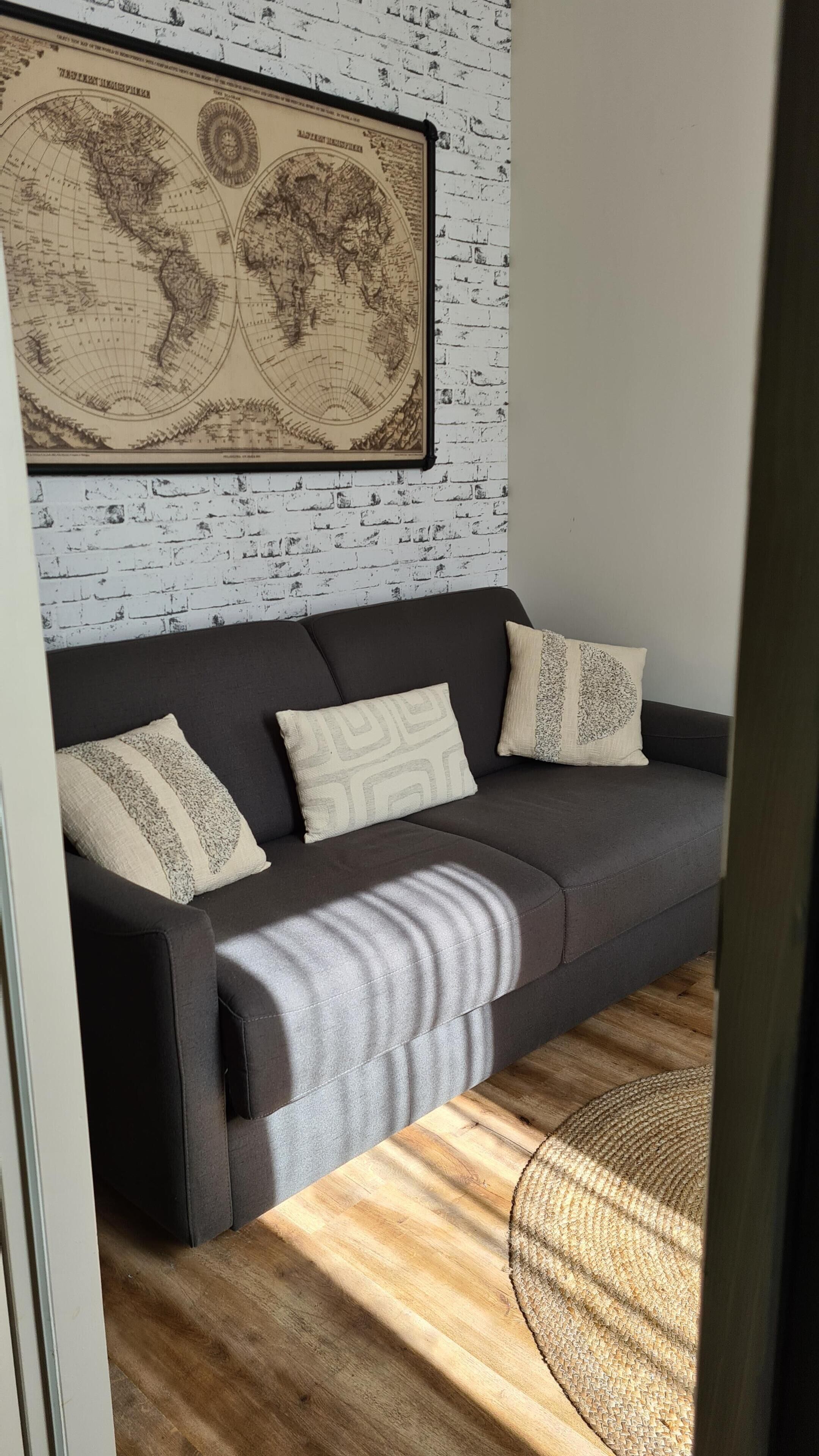 A dark grey sofa with patterned cushions against a white brick wall, a world map above, and a woven floor lamp.