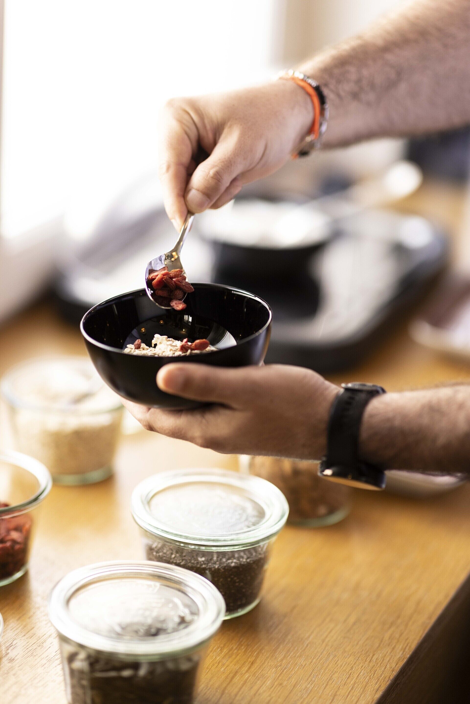 A person holding a bowl with a spoon, with various jars of ingredients on a wooden surface.
