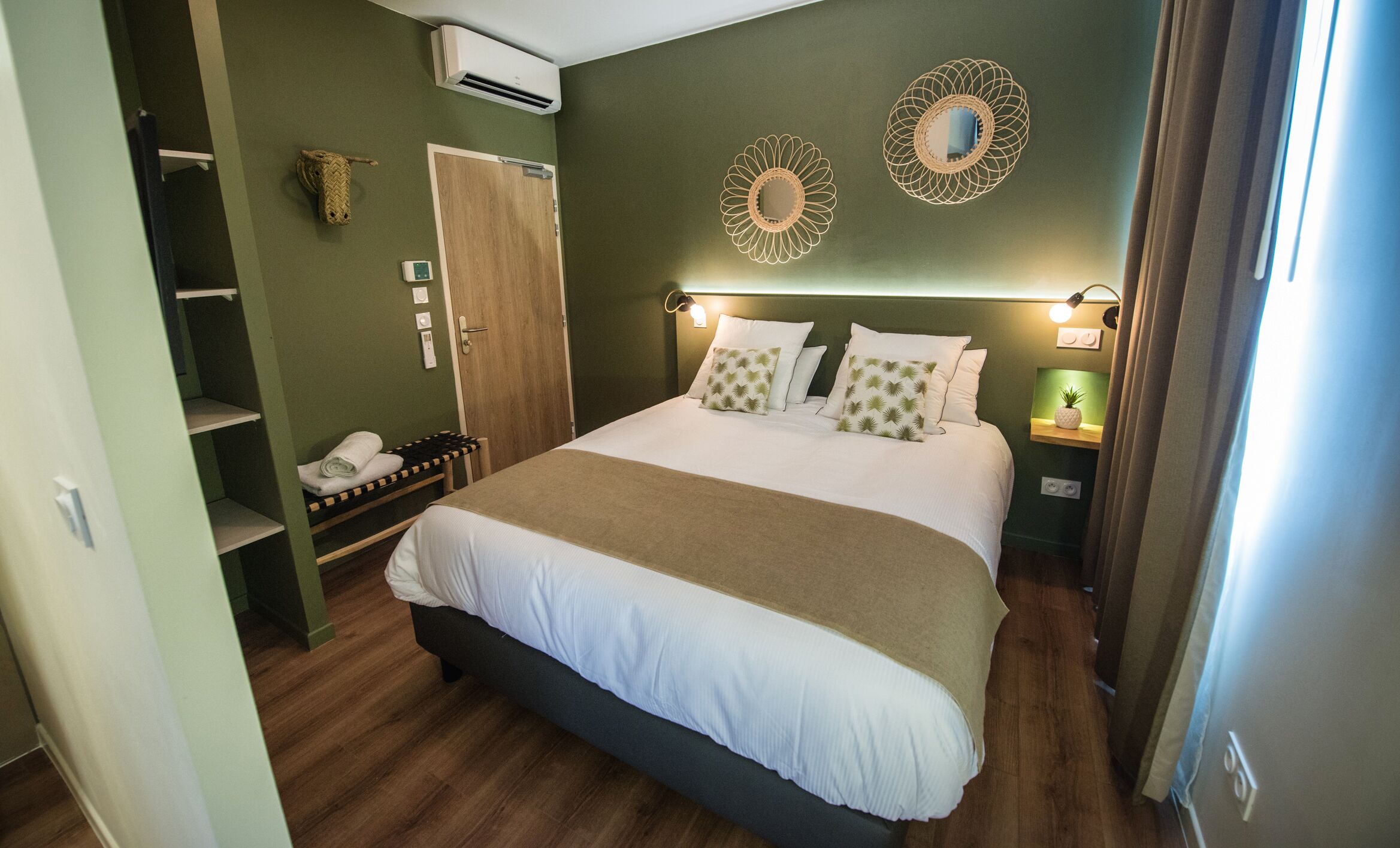 A hotel room with a large bed, a wooden floor, a wardrobe, a shelf with towels, and two wall-mounted decorative mirrors.