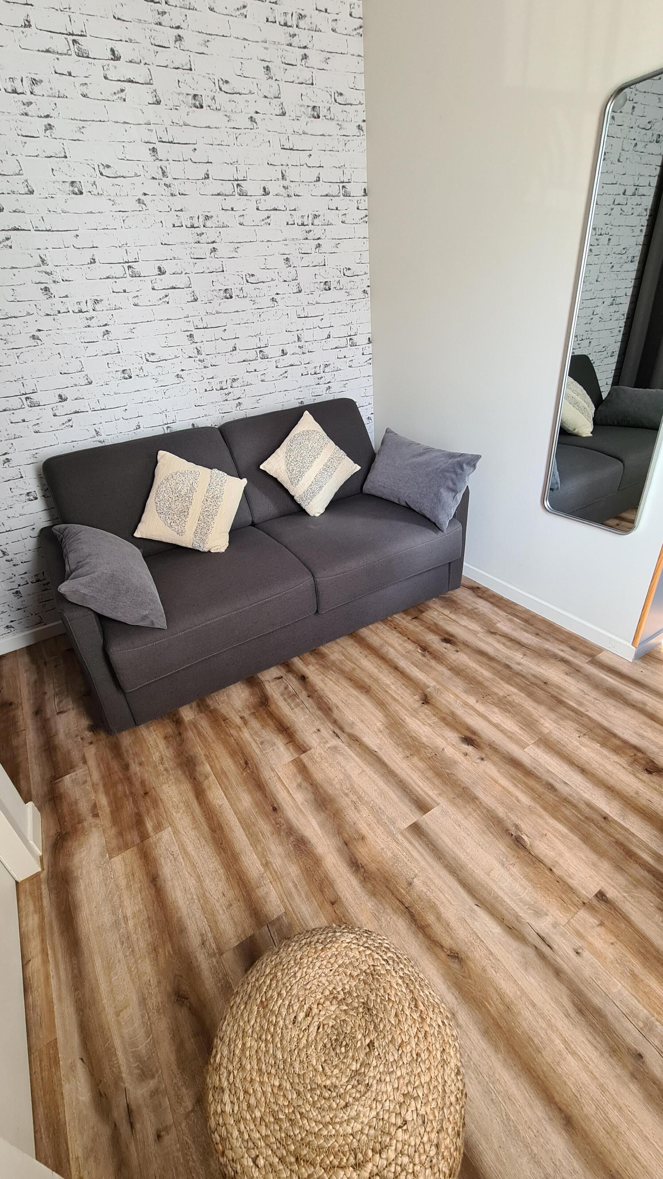Una sala de estar moderna con un sofá gris oscuro, cojines estampados, un taburete tejido y un espejo que refleja la habitación.