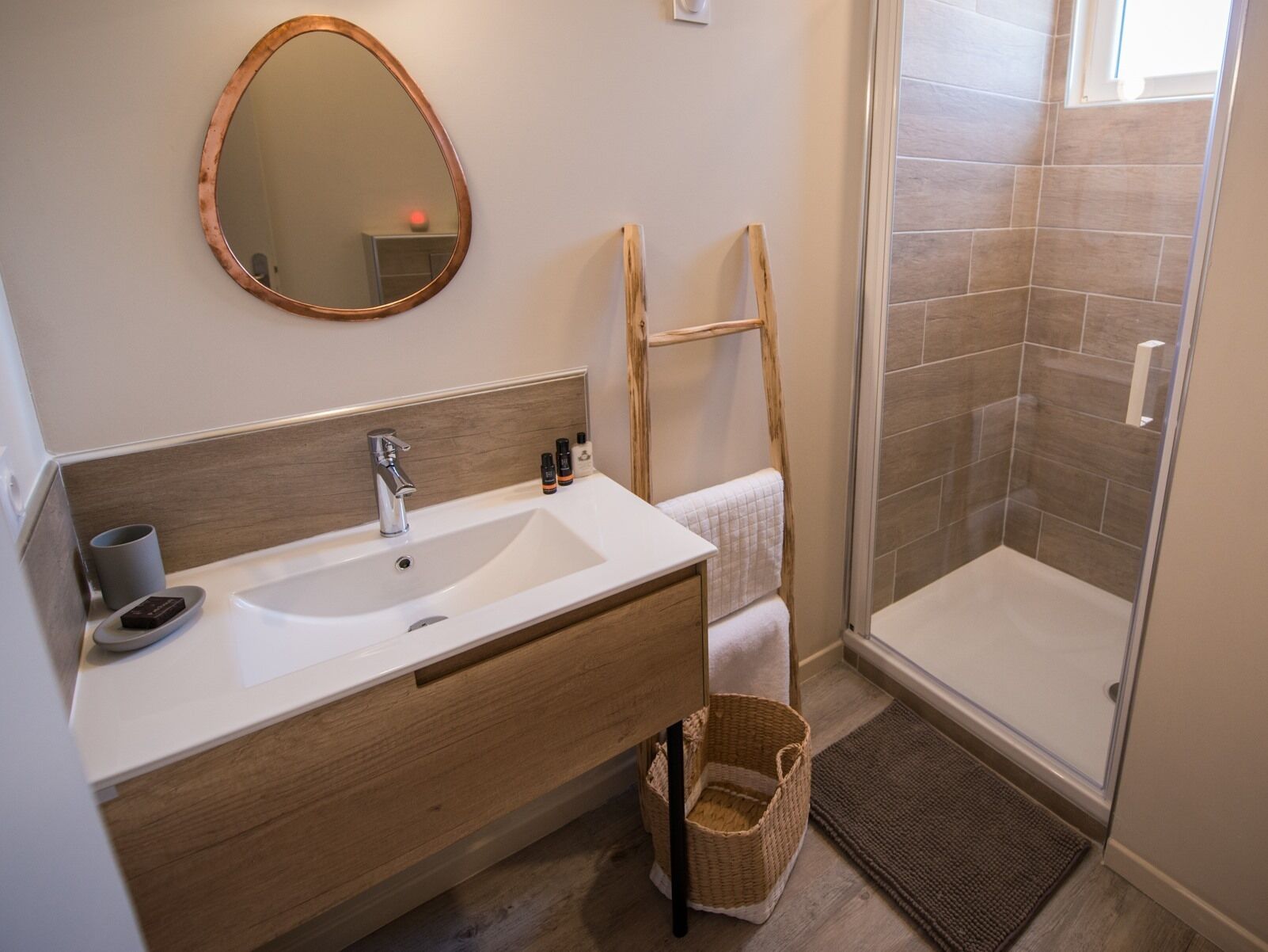 Un baño con lavamanos blanco, tocador de madera, espejo, toallero, ducha con paredes de azulejos y una cesta de mimbre.