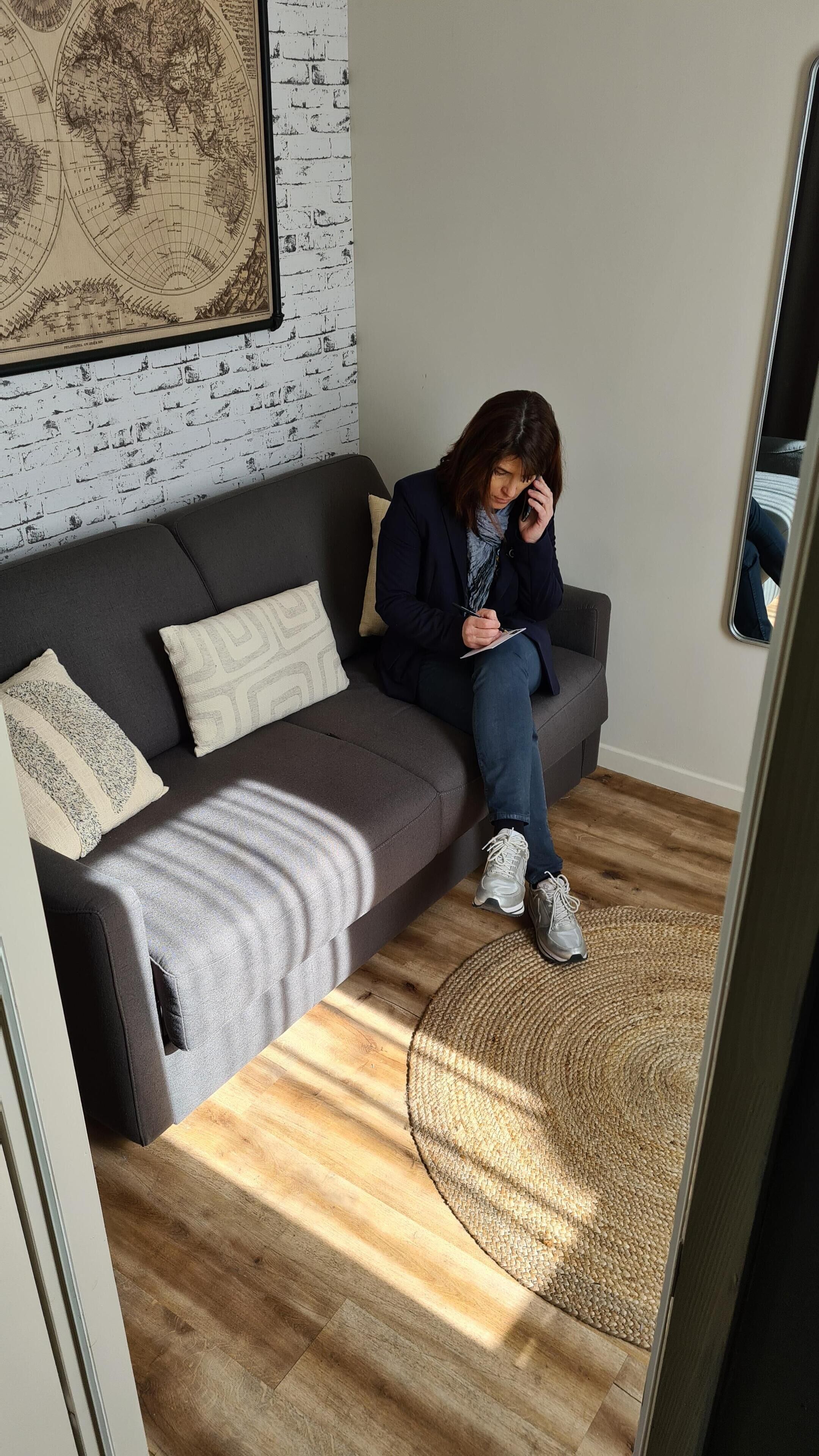 A person sitting on a grey sofa with patterned cushions, a world map on the wall, and a woven rug on the wooden floor.