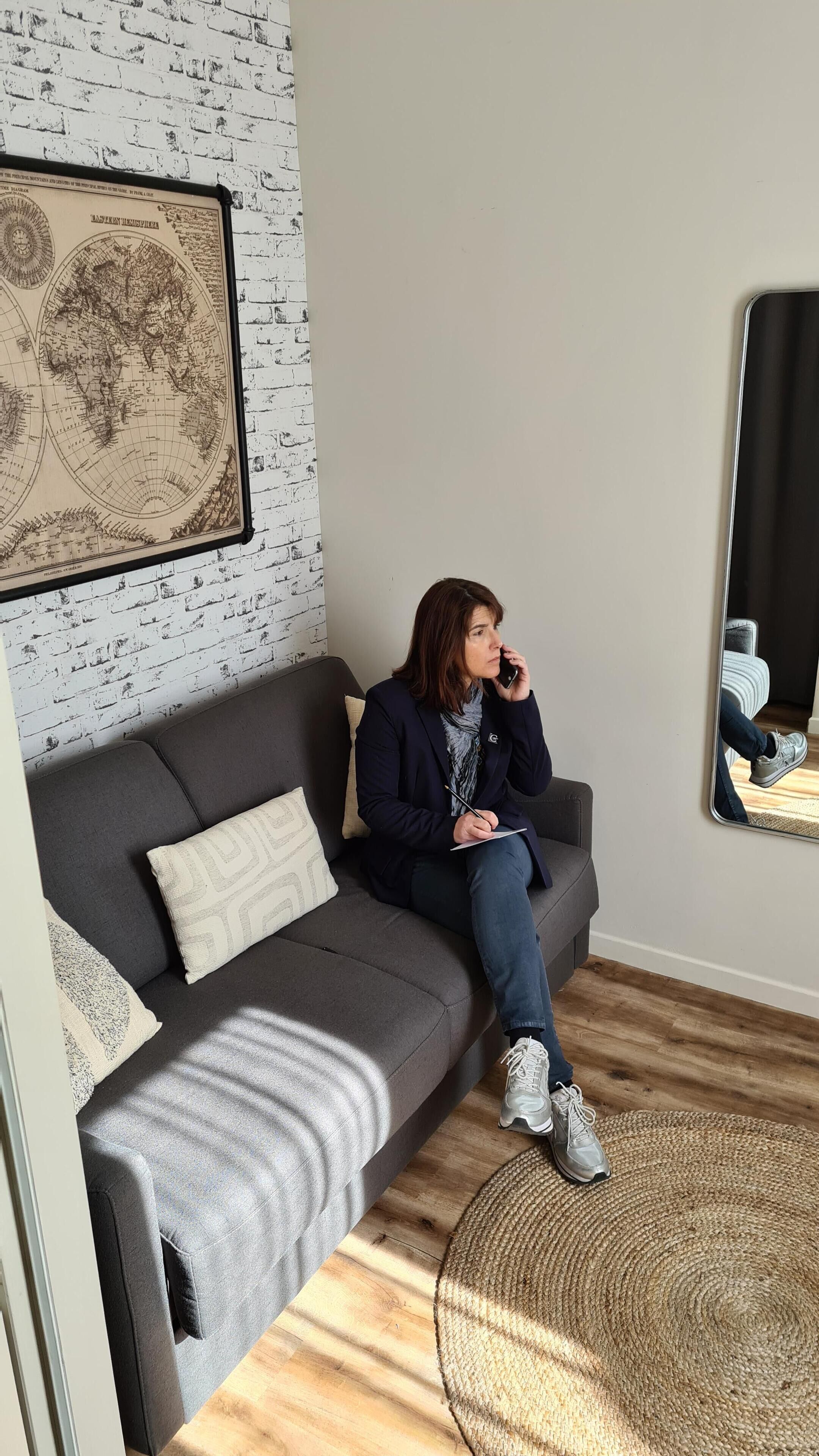 A person sitting on a grey sofa with a world map on the wall behind them, a woven rug on the floor, and a mirror reflecting part of the room.