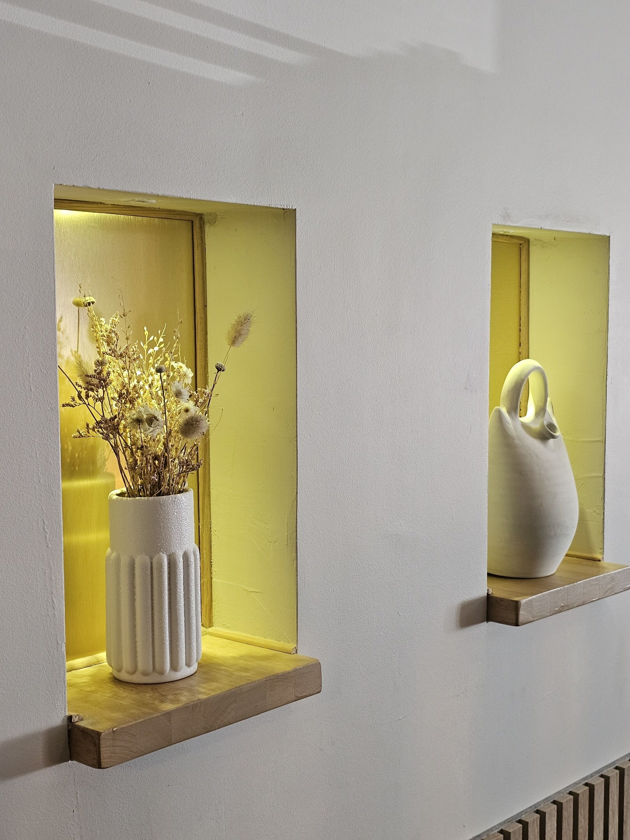 Deux vases blancs ornés de fleurs séchées sont posés sur des étagères, le tout adossé à un mur.