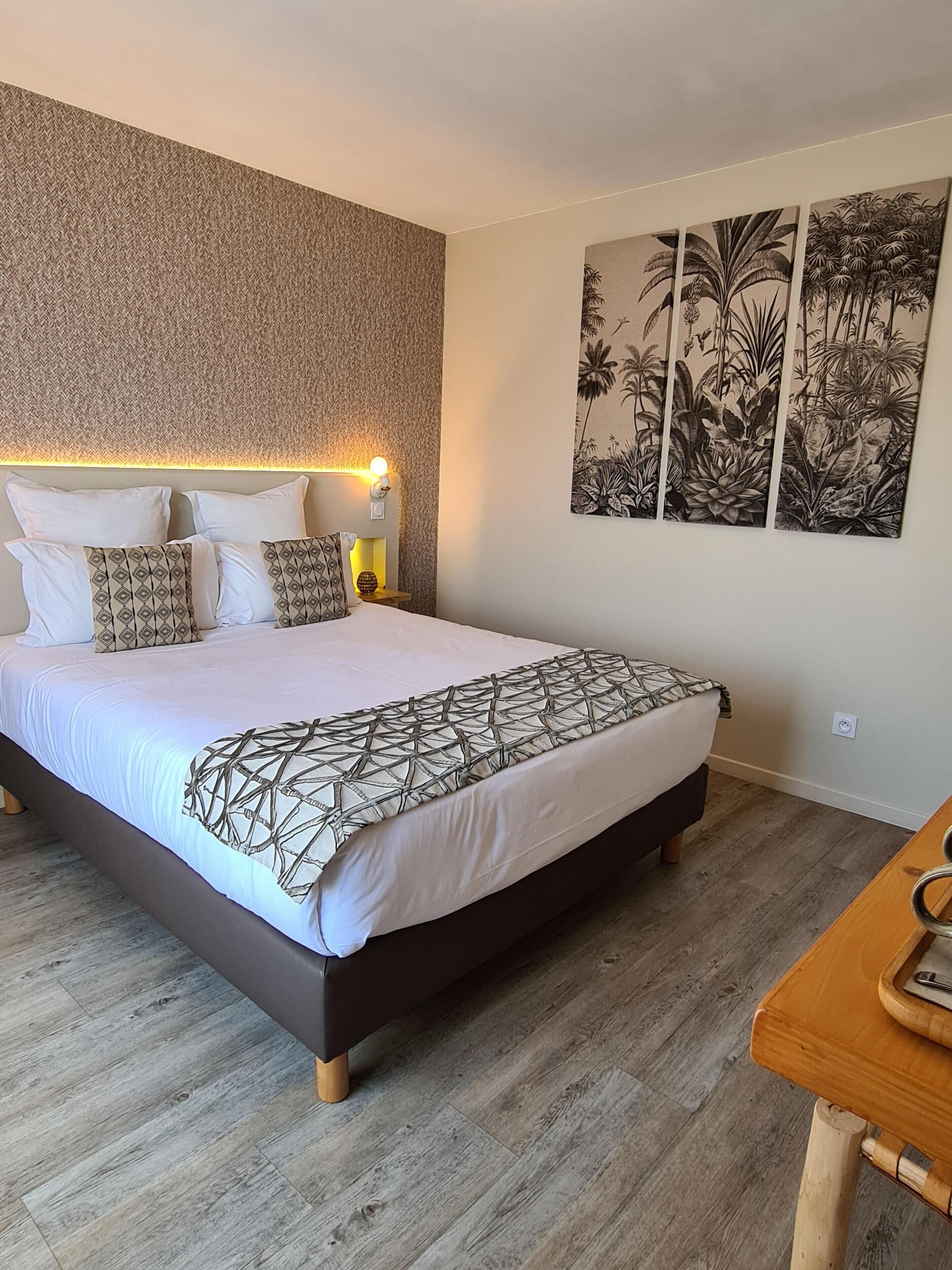 A bedroom with a large bed, a wooden nightstand, a wall-mounted lamp, and a large botanical print on the wall.