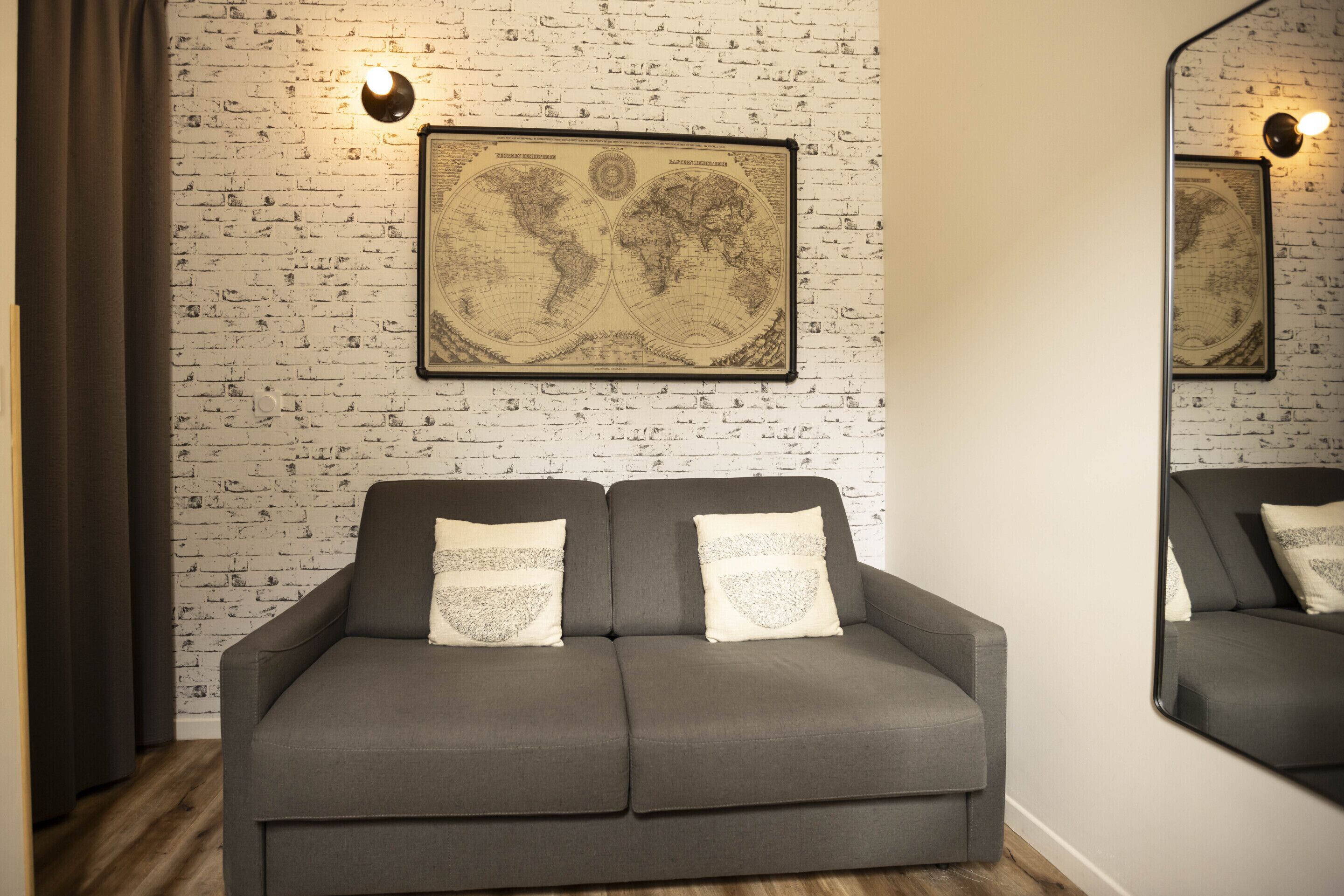 Un canapé gris agrémenté de coussins décoratifs, placé devant un mur dont le motif rappelle celui d’un mur de briques, et sur lequel est accrochée une carte du monde encadrée.