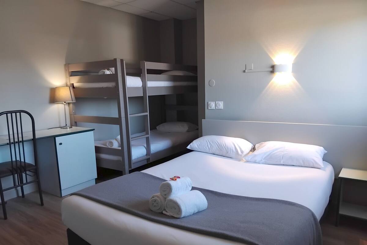 A hotel room with bunk beds, a desk with a lamp, a chair, and a neatly made bed with rolled towels.