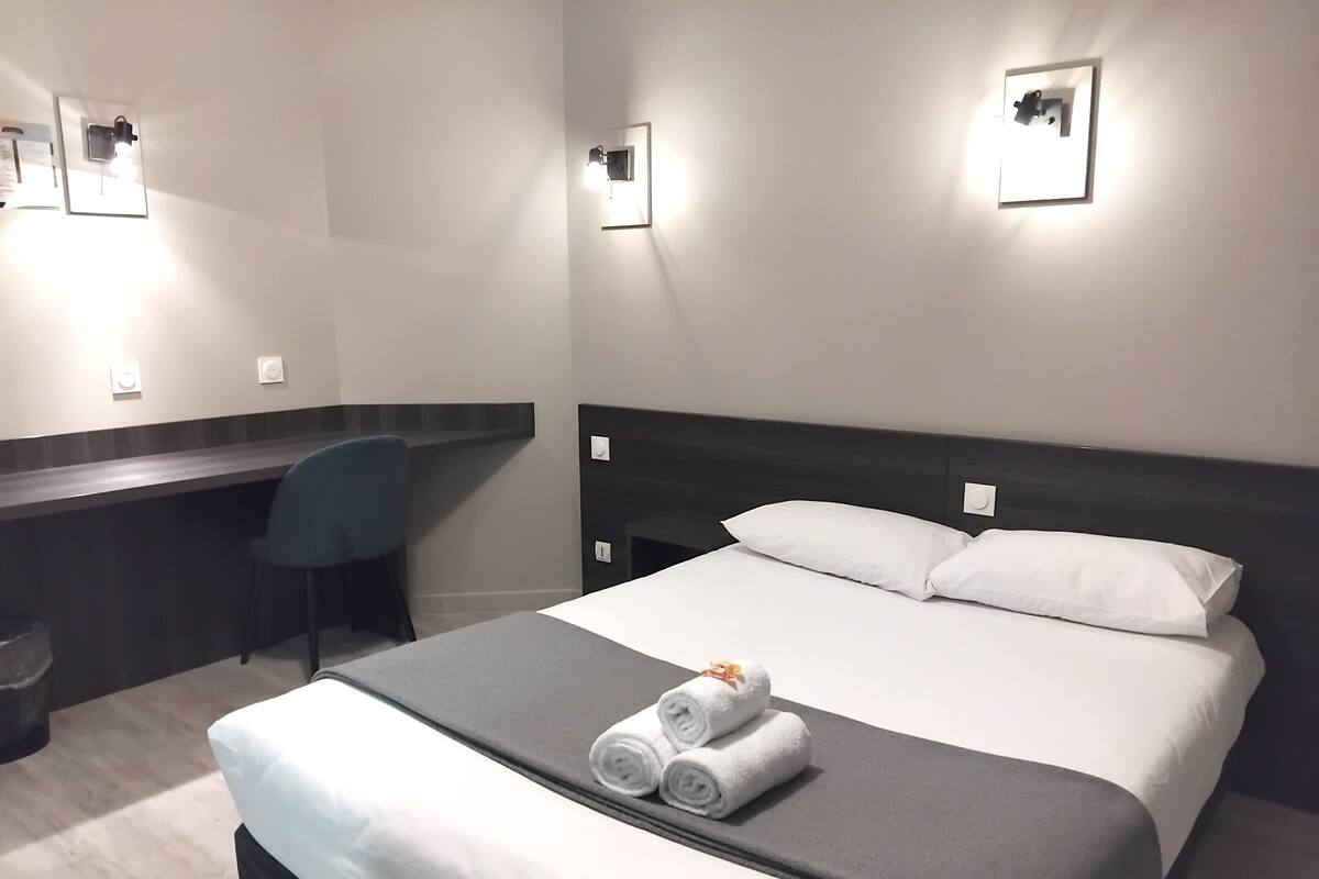 A hotel room with a bed, a desk, and a chair. There are two wall-mounted lights and a picture on the wall.