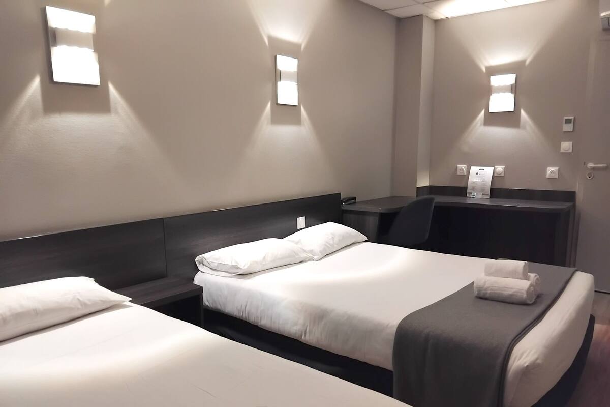 A hotel room with two beds, a desk, and a chair. There are wall-mounted lights and a thermostat.