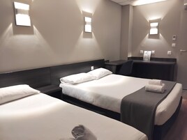 Quarto de hotel com duas camas, uma escrivaninha e uma cadeira. Há luminárias fixadas na parede e um termostato.