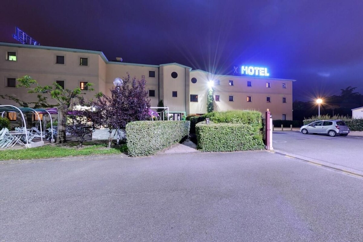 A hotel with a well-lit entrance, a parking area, and neatly trimmed hedges.