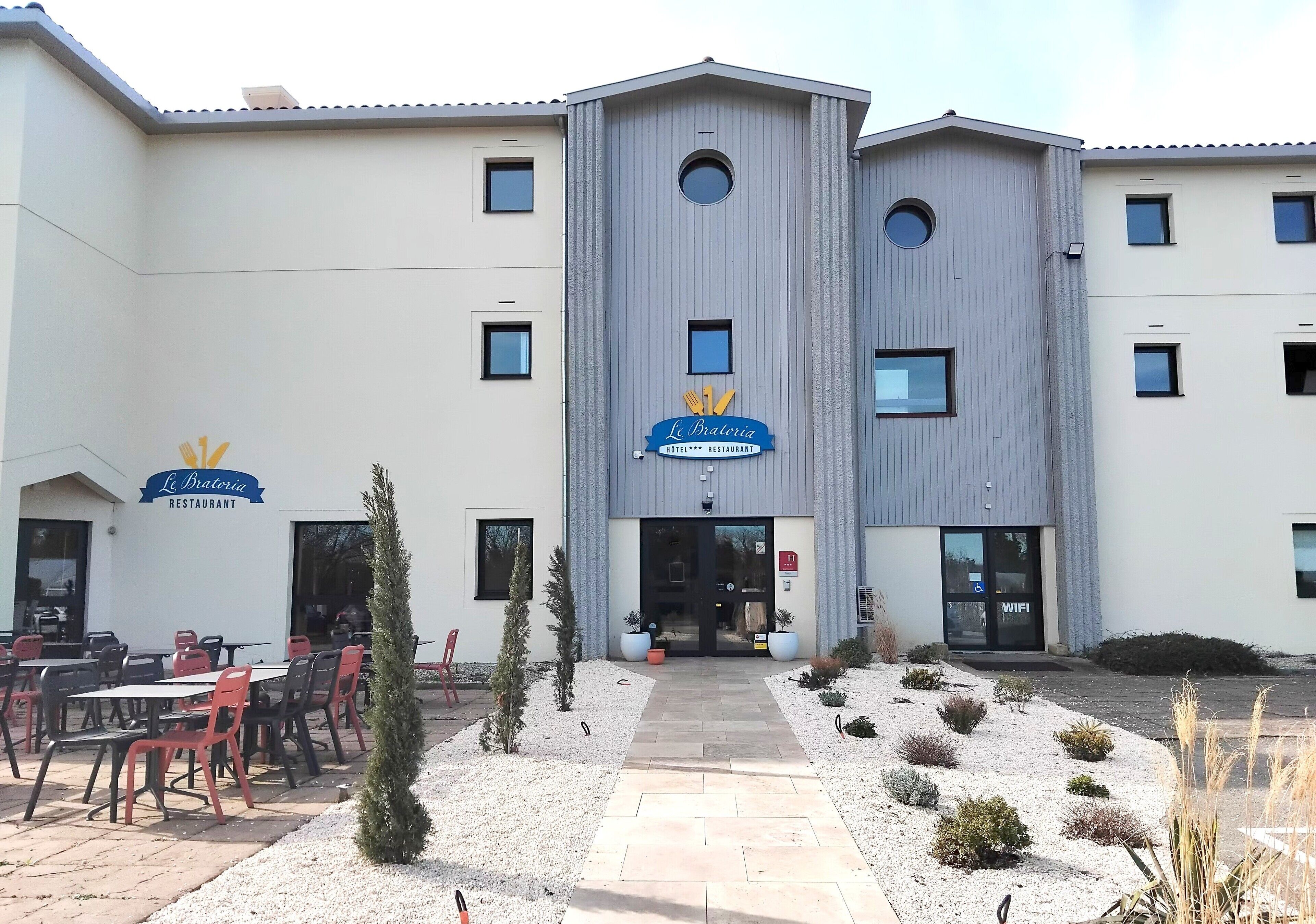 Un bâtiment moderne avec une enseigne indiquant « La Bateria Restaurant » et une autre indiquant « WiFi ». On y trouve des tables et des chaises à l’extérieur, ainsi qu’un chemin menant à l’entrée.