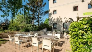 En udendørs terrasse med borde og stole, omgivet af grøn vegetation og en bygning med et skilt.