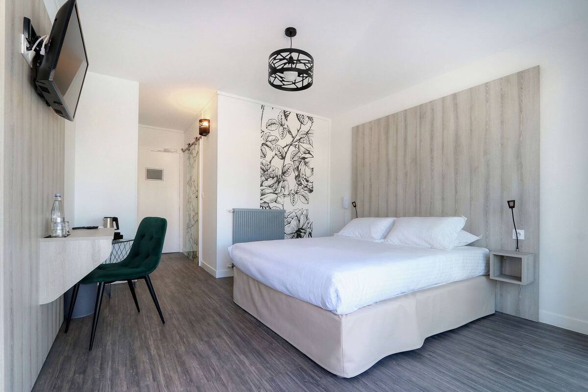 Quarto de hotel com cama, escrivaninha com cadeira, televisão fixada na parede e radiador.
